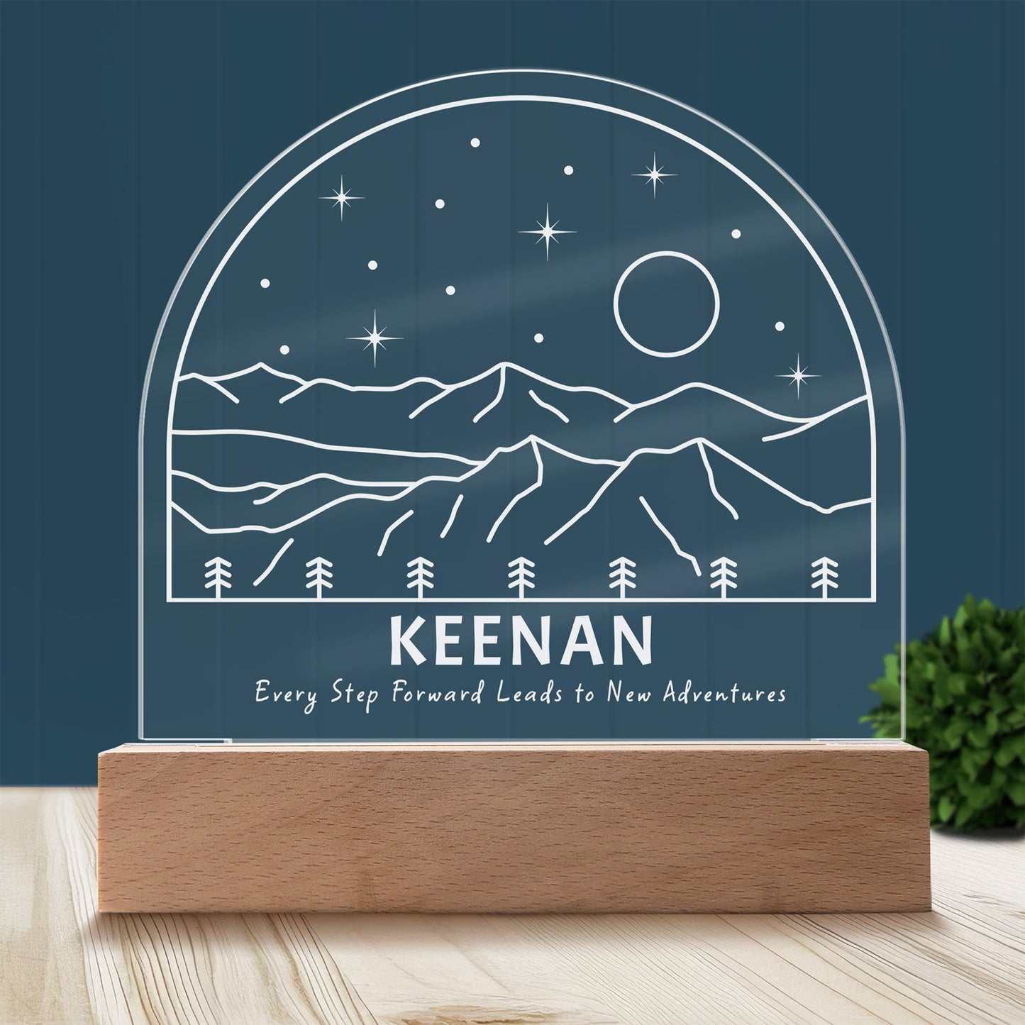 New Adventures Personalized Night Light with Battery powered base featured on a light timber table with a dark blue panelled wall and lush green pot plant in the background.