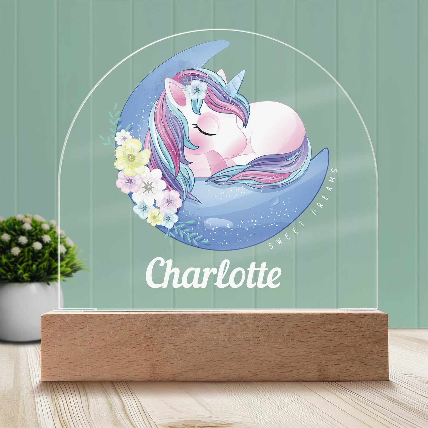 Sweet Dreams Unicorn Personalized Night Light with Battery powered base featured on a light timber table with a mint green panelled wall and lush green pot plant with white flowers in the background.