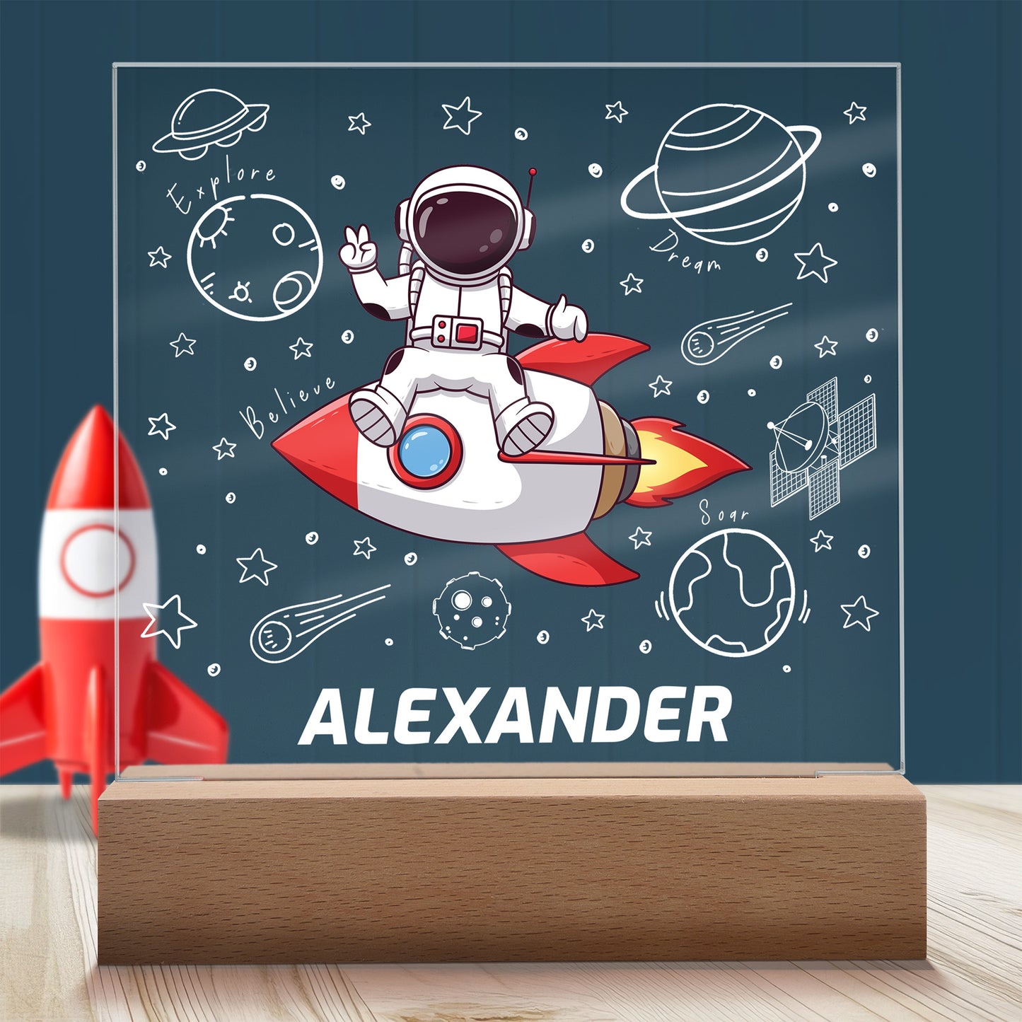 Astronaut on a Rocket Personalized Night Light with Battery powered base featured on a light timber table with a dark blue panelled wall and child's rocket toy in the background.