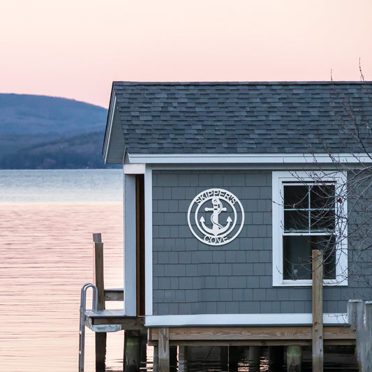 Elaborate Anchor Ring Metal Decor in White with the text Skippers Cove is installed on the blue shingle wall of a boat shed on a lake.