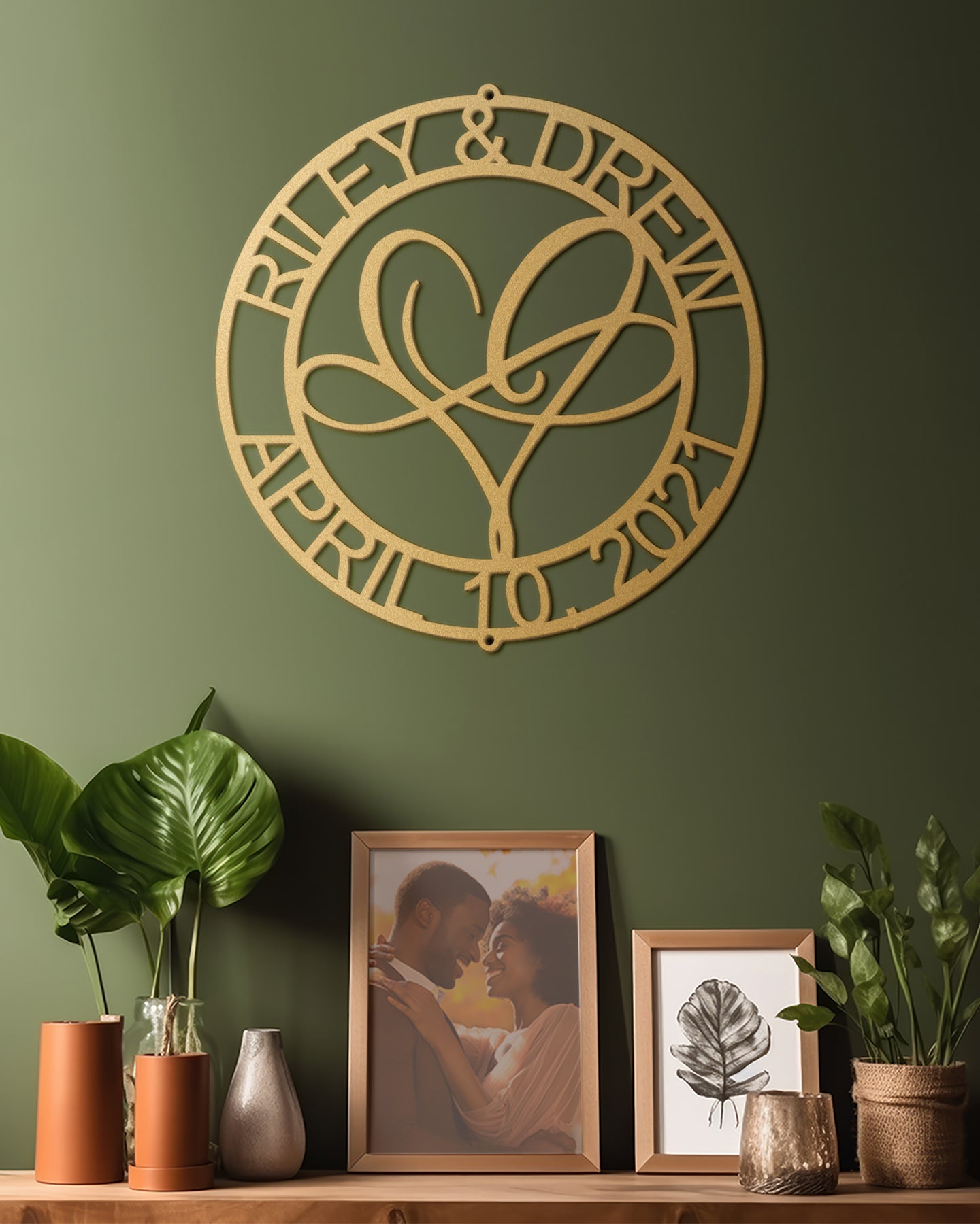 Infinite Love Personalized Metal Decor in Gold customized with names and date on a green wall, below which are framed photos and plants.