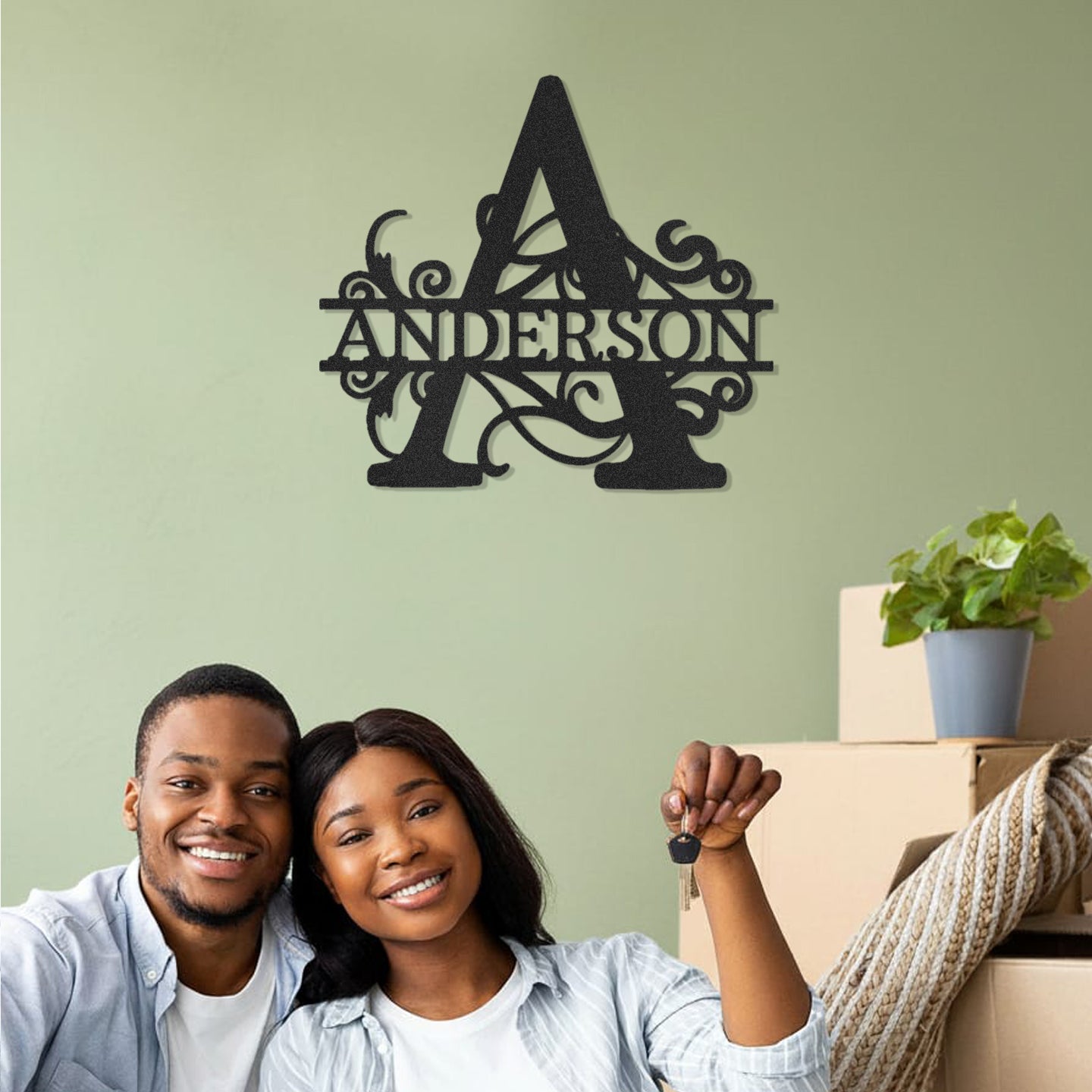 Couple sitting together with moving boxes, smiling at the camera, against a light green wall with a Split Letter Monogram Personalized Metal Decor piece mounted above them.