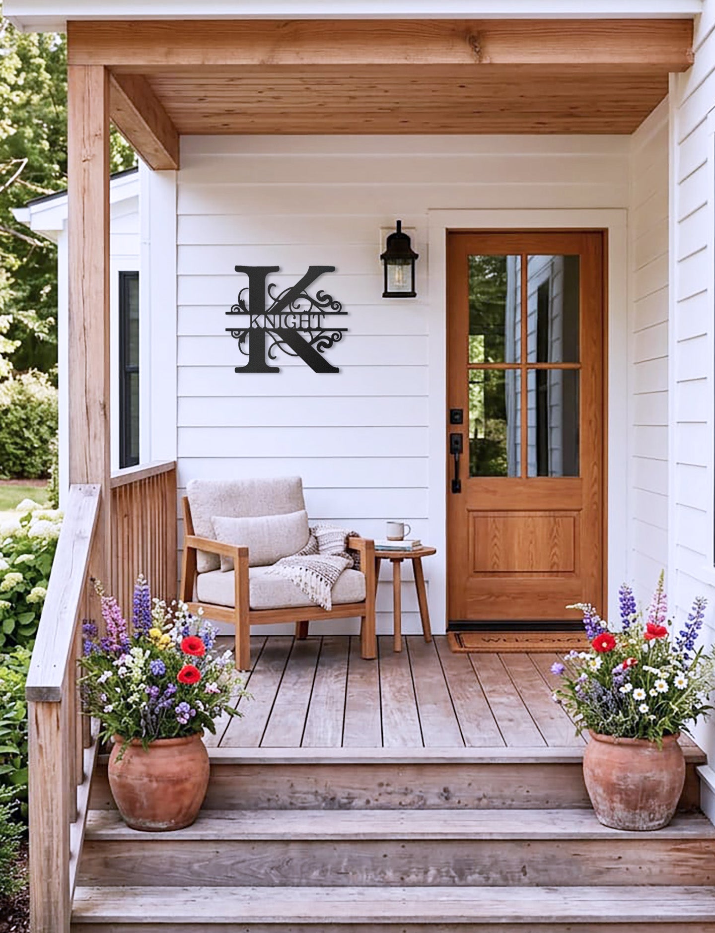Black Split Letter Monogram Personalized Metal Decor piece mounted on the wall of the front porch of a white country style house, complemented by a wooden chair, table, and potted plants.