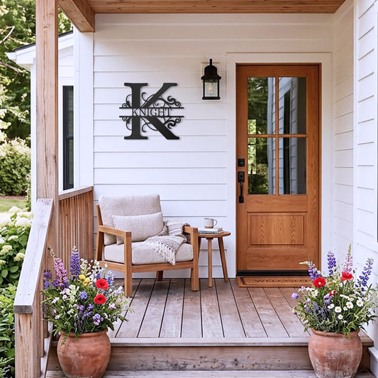 Black Split Letter Monogram Personalized Metal Decor piece mounted on the wall of the front porch of a white country style house, complemented by a wooden chair, table, and potted plants.