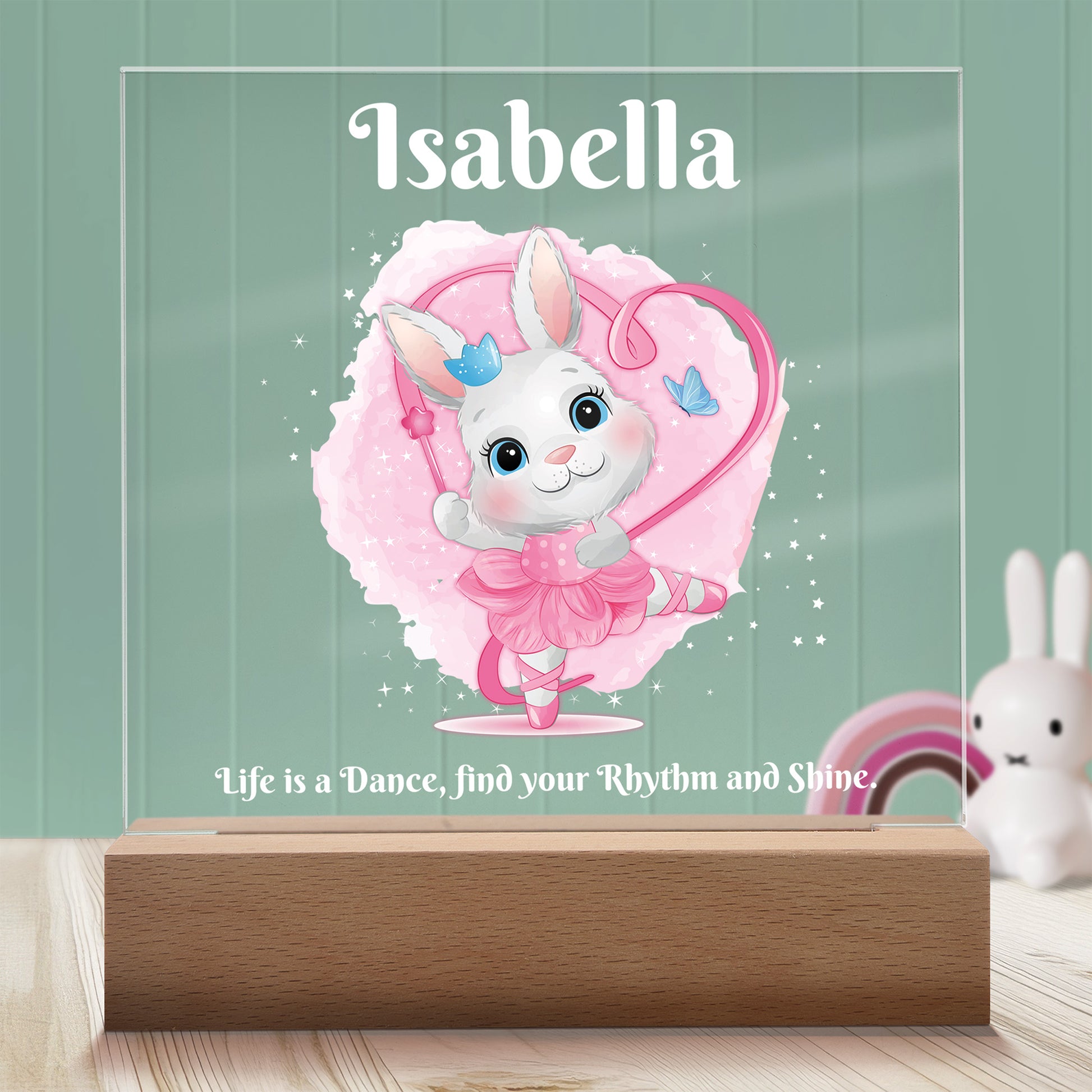 Dancing Bunny Personalized Night Light with Battery powered base featured on a light timber table with a mint green panelled wall and bunny and rainbow ornaments in the background.