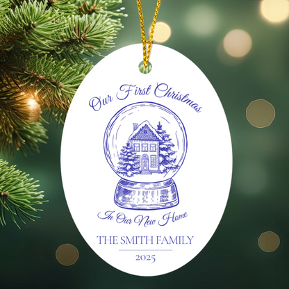 Decorative ornament with 'Our First Christmas in Our New Home' text and a house illustration, against a blurred Christmas tree background.