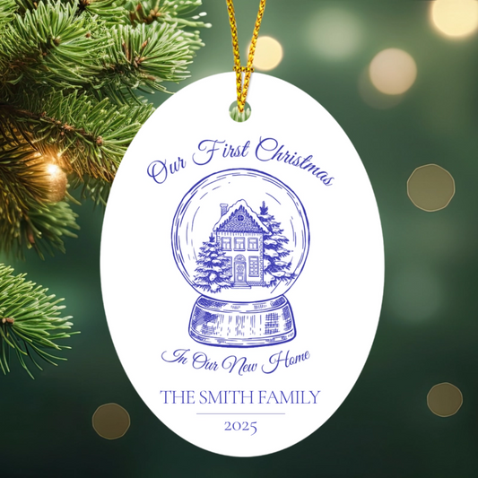 Decorative ornament with 'Our First Christmas in Our New Home' text and a house illustration, against a blurred Christmas tree background.