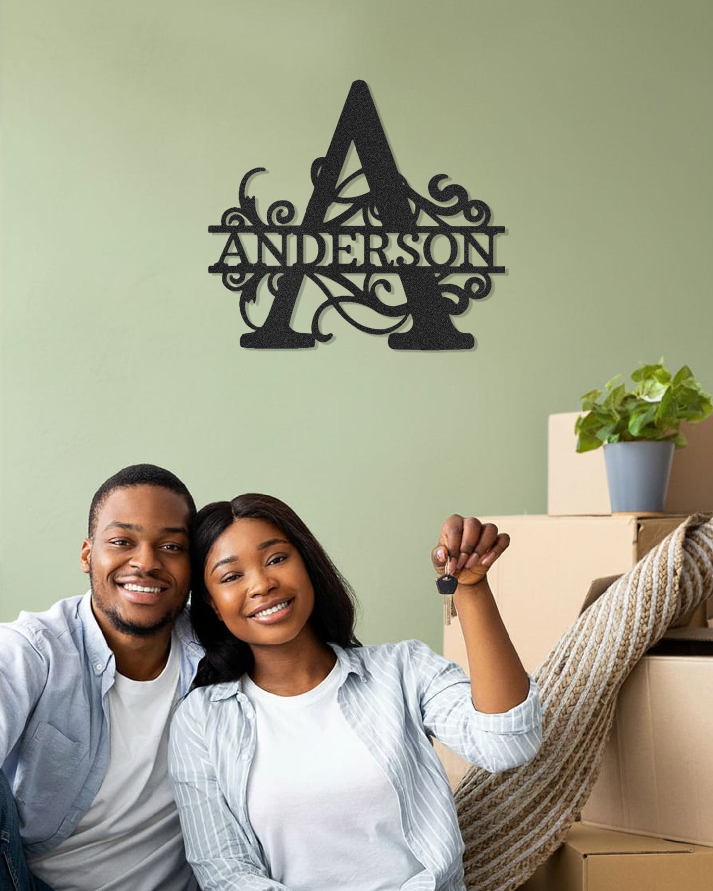 Couple sitting together with moving boxes, smiling at the camera, against a light green wall with a Split Letter Monogram Personalized Metal Decor piece mounted above them.