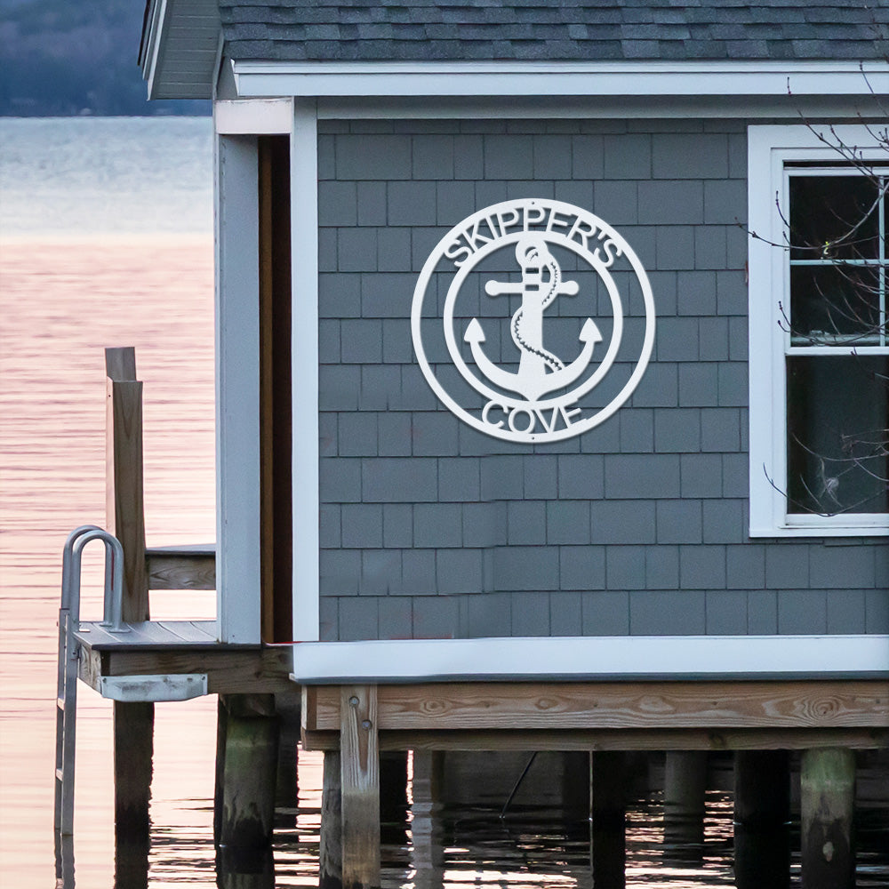 Elaborate Anchor Ring Metal Decor in White with the text Skippers Cove is installed on the blue shingle wall of a boat shed.